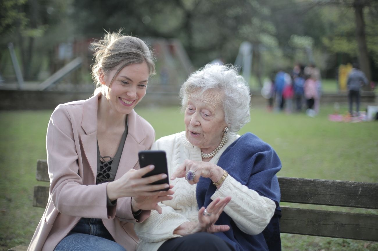 Young and older woman sit on a bench, looking at our services on a phone screen.