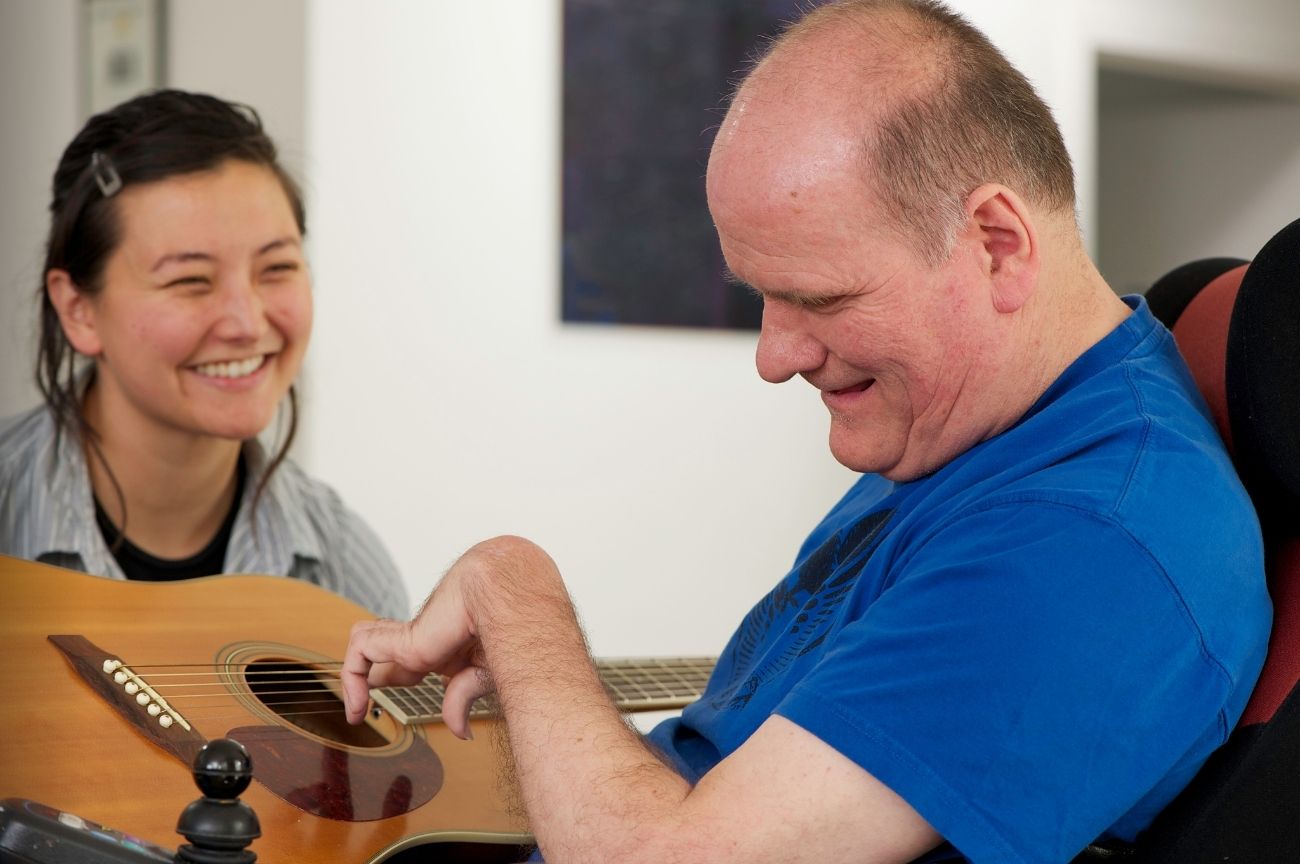 A disabled man plays guitar with support from his Personal Assistant.
