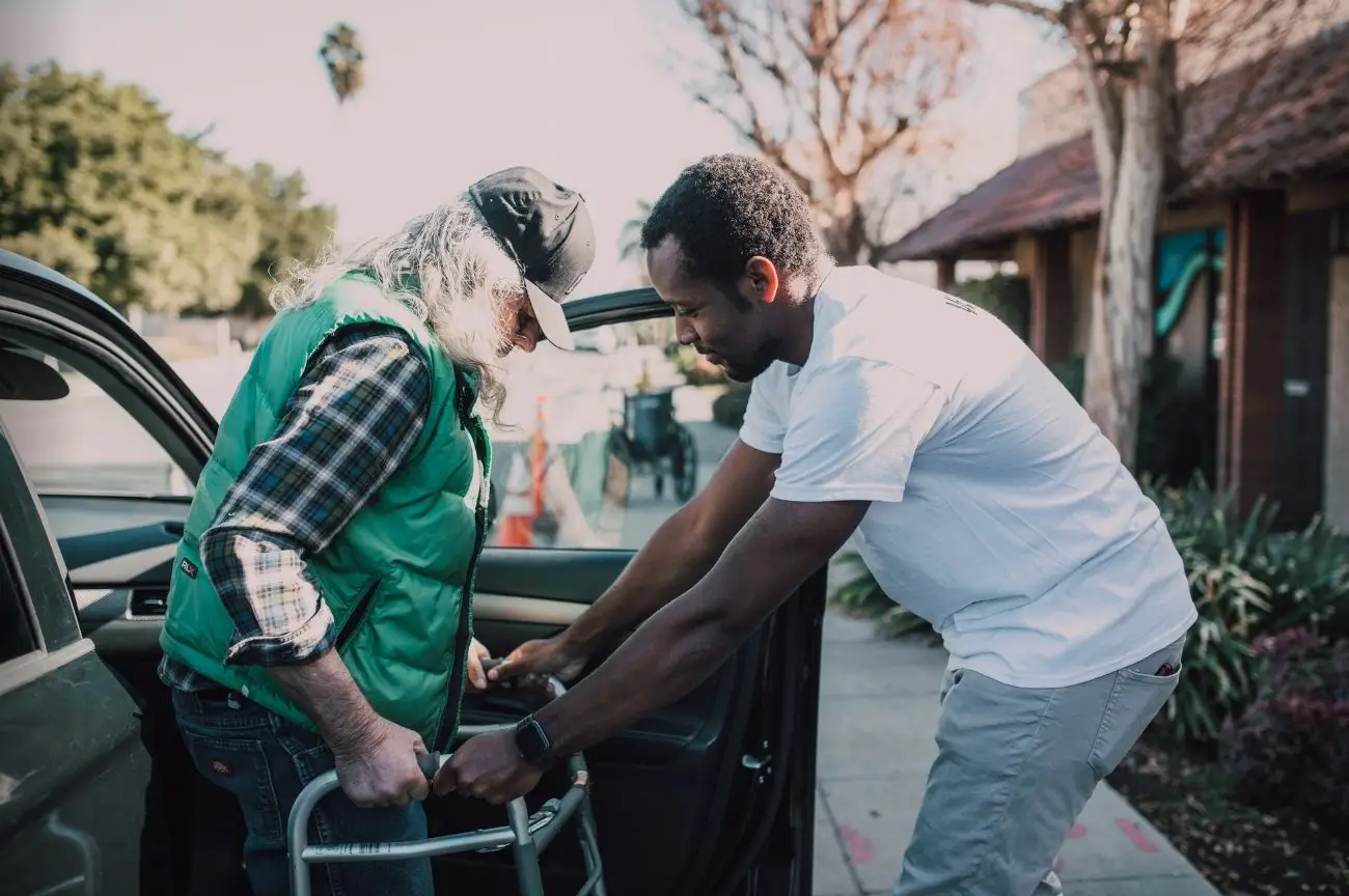 A man helps another man with a walking frame to get out of a car, showing support for self-funders.